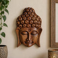 A carved wooden Buddha head wall sculpture in a warm medium brown tone, mounted on a beige wall. A green houseplant is visible on the left, and the corner of a wooden picture frame and a decorative lamp are visible on the right.