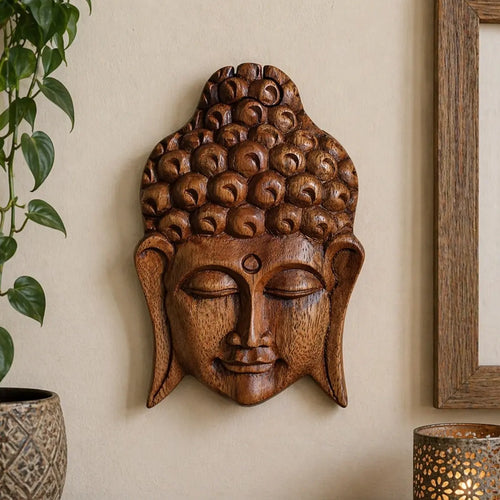 A carved wooden Buddha head wall sculpture in a warm medium brown tone, mounted on a beige wall. A green houseplant is visible on the left, and the corner of a wooden picture frame and a decorative lamp are visible on the right.