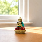 A small decorative Buddha statue seated on a lotus base, placed on a wooden table near a window with soft natural light. The statue has a gold-toned face, hands, and base, with turquoise and red mosaic details on the robe and seat.