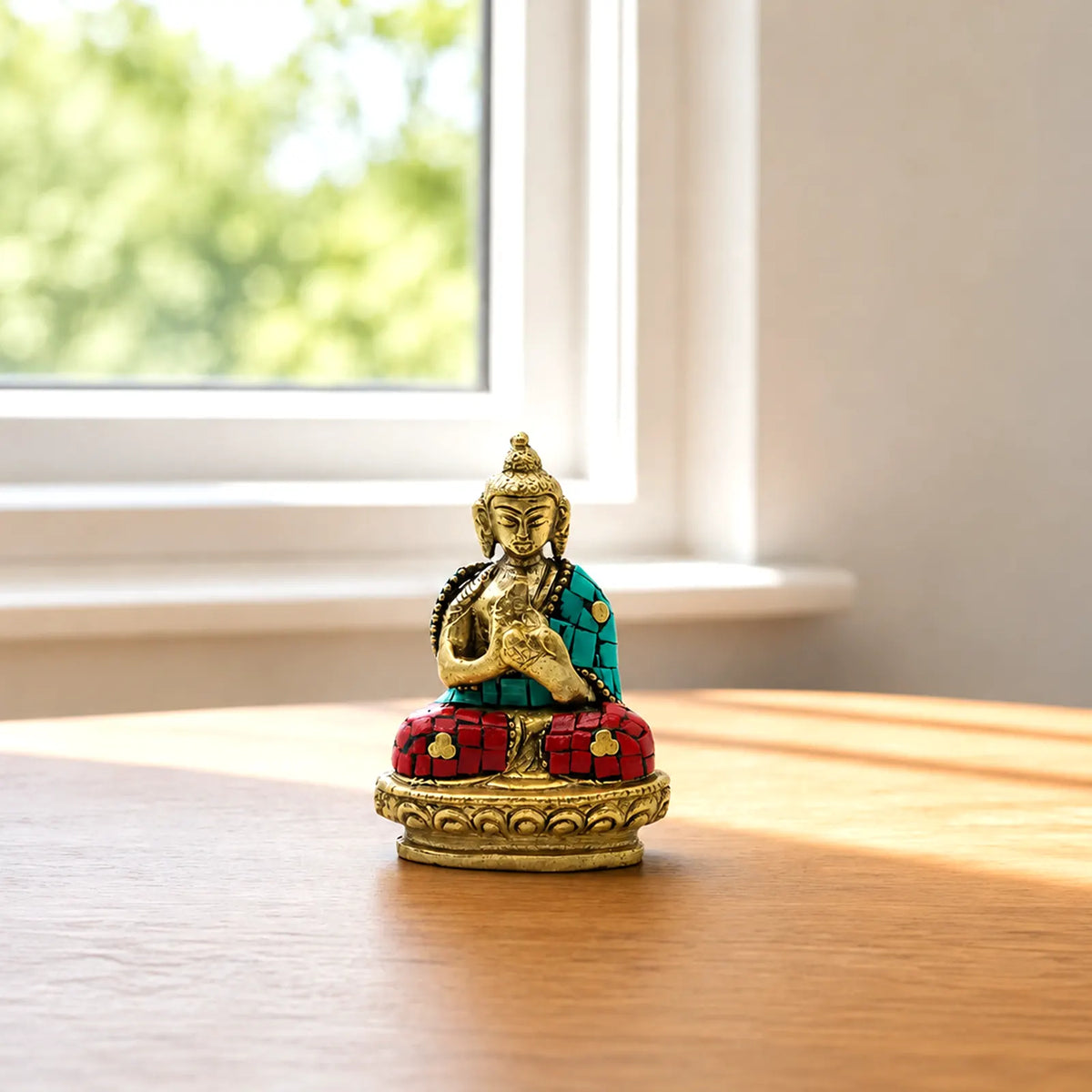 A small decorative Buddha statue seated on a lotus base, placed on a wooden table near a window with soft natural light. The statue has a gold-toned face, hands, and base, with turquoise and red mosaic details on the robe and seat.