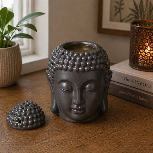 Buddah Head Oil Burner - Dark Brown, a detailed Buddha head design oil burner in dark brown, placed on a wooden surface next to a lit decorative candle and books for peaceful home decor.