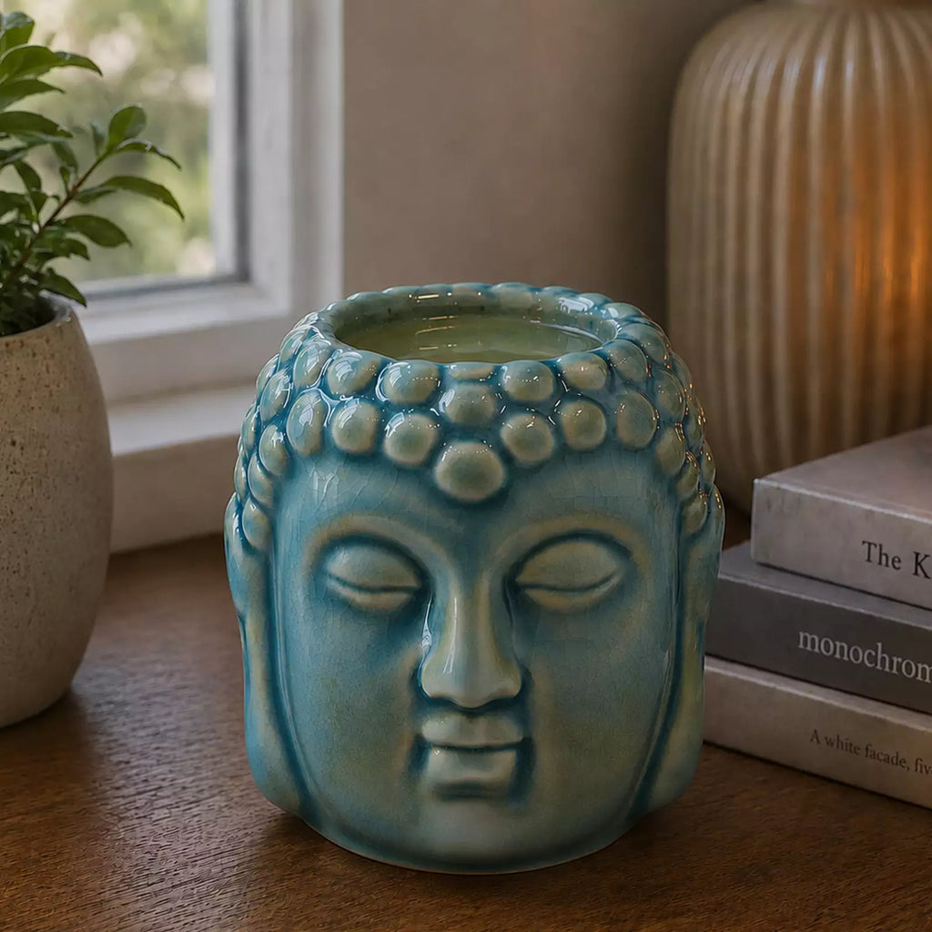 Buddha Oil Burner - Blue featuring a serene Buddha face design with a lit candle inside, placed on a wooden surface beside books and decorative items.