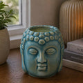 Buddha Oil Burner - Blue featuring a serene Buddha face design with a lit candle inside, placed on a wooden surface beside books and decorative items.