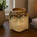 Buddha Oil Burner - Ivory & Mint ceramic oil burner shaped like a Buddha head with a lit flame, placed on a wooden surface near books and decorative candles.