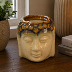 Buddha Oil Burner - Ivory & Mint ceramic oil burner shaped like a Buddha head with a lit flame, placed on a wooden surface near books and decorative candles.