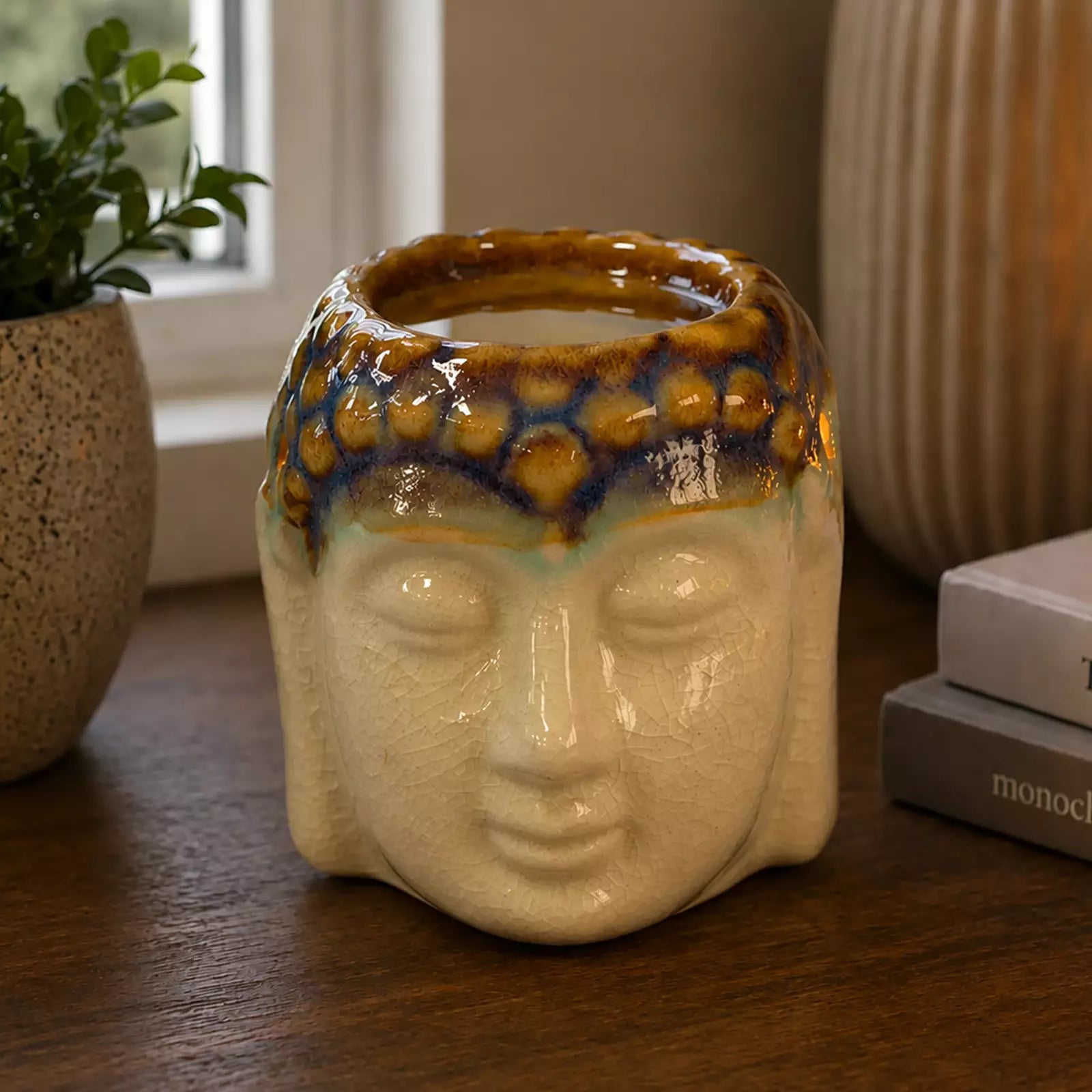 Buddha Oil Burner - Ivory & Mint ceramic oil burner shaped like a Buddha head with a lit flame, placed on a wooden surface near books and decorative candles.
