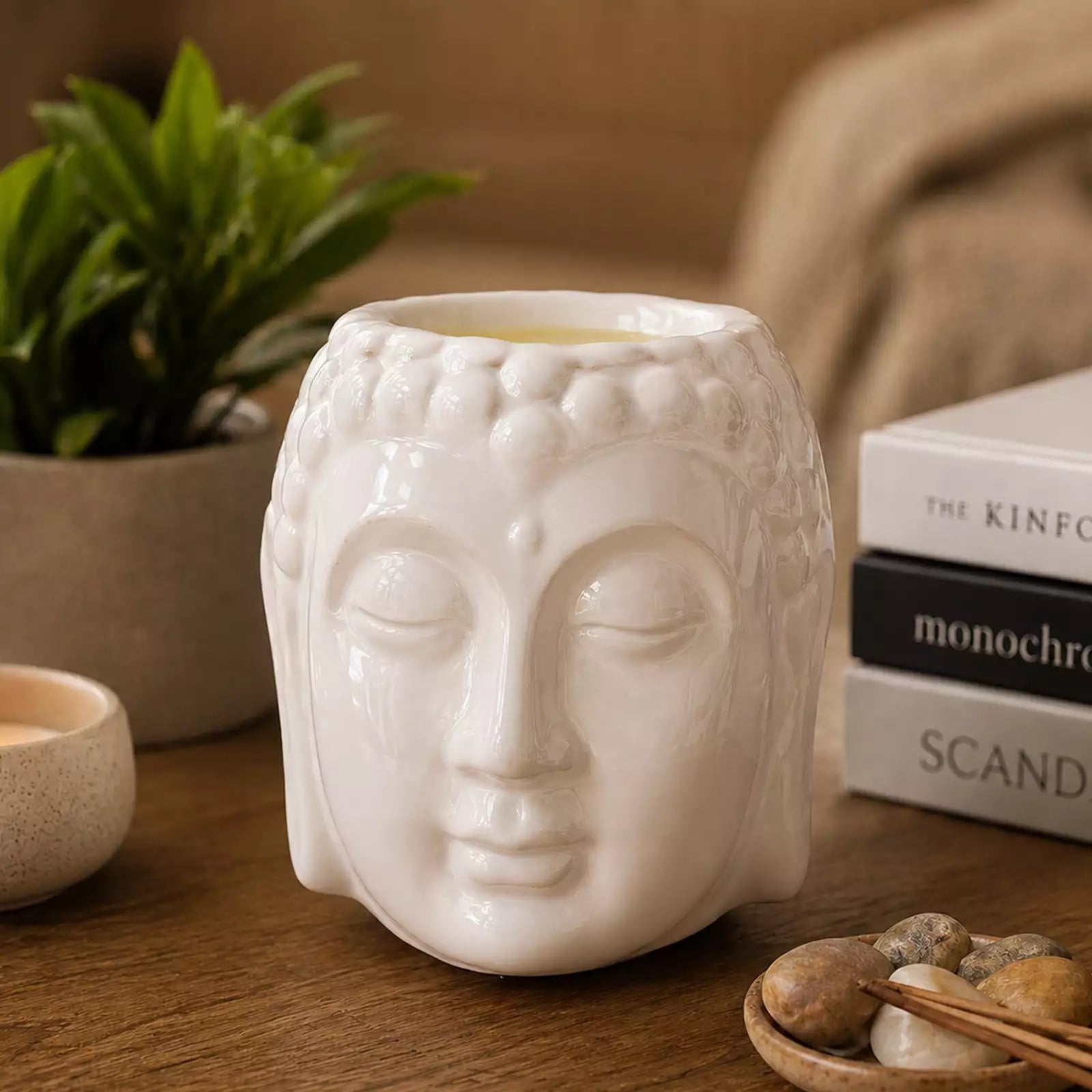 Buddha Oil Burner - White with a serene Buddha face design releasing fragrant steam, placed on a wooden surface surrounded by calming décor including a plant and books.
