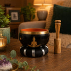 Buddha Singing Bowl Set- Black/Orange 10.7cm featuring a black bowl with gold Buddha silhouette on a striped cushion and a wooden striker, displayed on a wooden table in a cozy room.