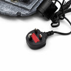A close-up of a black UK three-pin power plug with a red safety cover, attached to a black cord, with part of a gray textured product visible at the top on a white background.