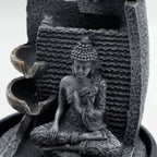 A dark gray stone Buddha figurine seated in a meditative pose, integrated into a tiered waterfall fountain structure with textured stone panels and curved water channels.