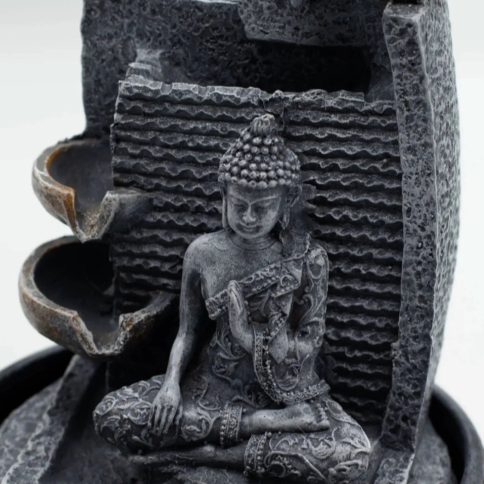 A dark gray stone Buddha figurine seated in a meditative pose, integrated into a tiered waterfall fountain structure with textured stone panels and curved water channels.