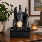 Buddha Hand & Head Tabletop Water Feature (20x25x35cm) with a serene Buddha head resting against a large hand, featuring a glowing orb and flowing water on a wooden table.