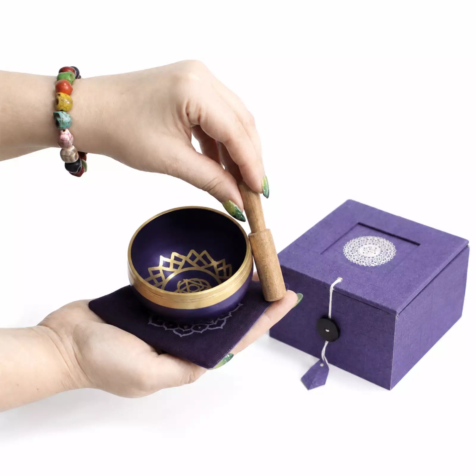 A purple singing bowl with a gold rim and a wooden mallet being held by hands over a matching purple cloth, with a purple gift box in the background.