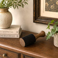 A wooden and felt floor roller lying on a dark wood table, styled in a cozy room setting with potted plants, a stack of books, and a framed picture in the background.