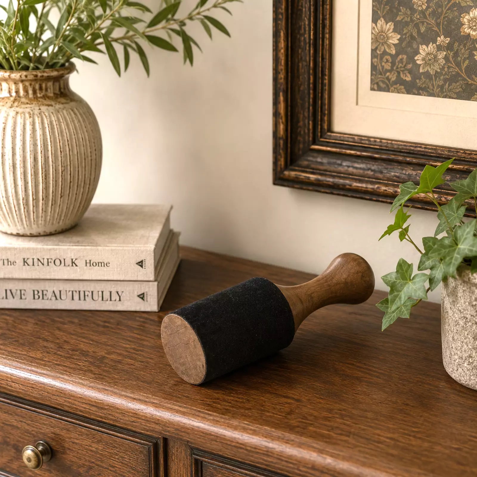 A wooden and felt floor roller lying on a dark wood table, styled in a cozy room setting with potted plants, a stack of books, and a framed picture in the background.