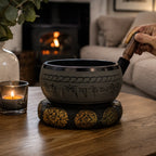 Extra Loud - Singing Bowl - Five Buddha resting on a decorative cushion on a wooden table next to a lit candle in a cozy room with a fireplace and soft seating in the background.