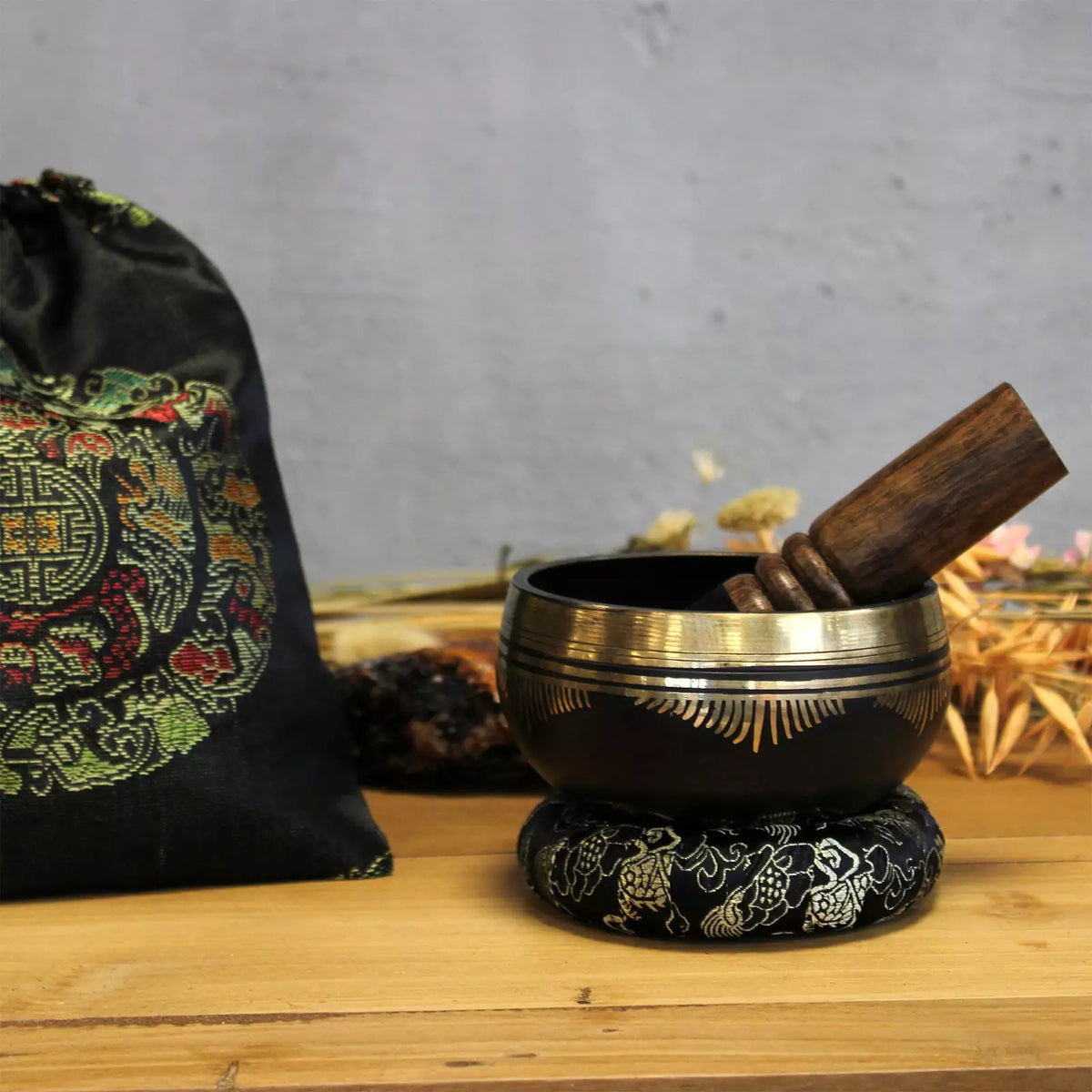 Decorative black and gold singing bowl with wooden striker on a wooden surface, with a patterned bag in the background.