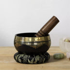 A black singing bowl with gold decorative patterns, resting on a matching black and gold decorative base, with a wooden mallet inside. The bowl is placed on a light wooden surface with a soft-focus background.