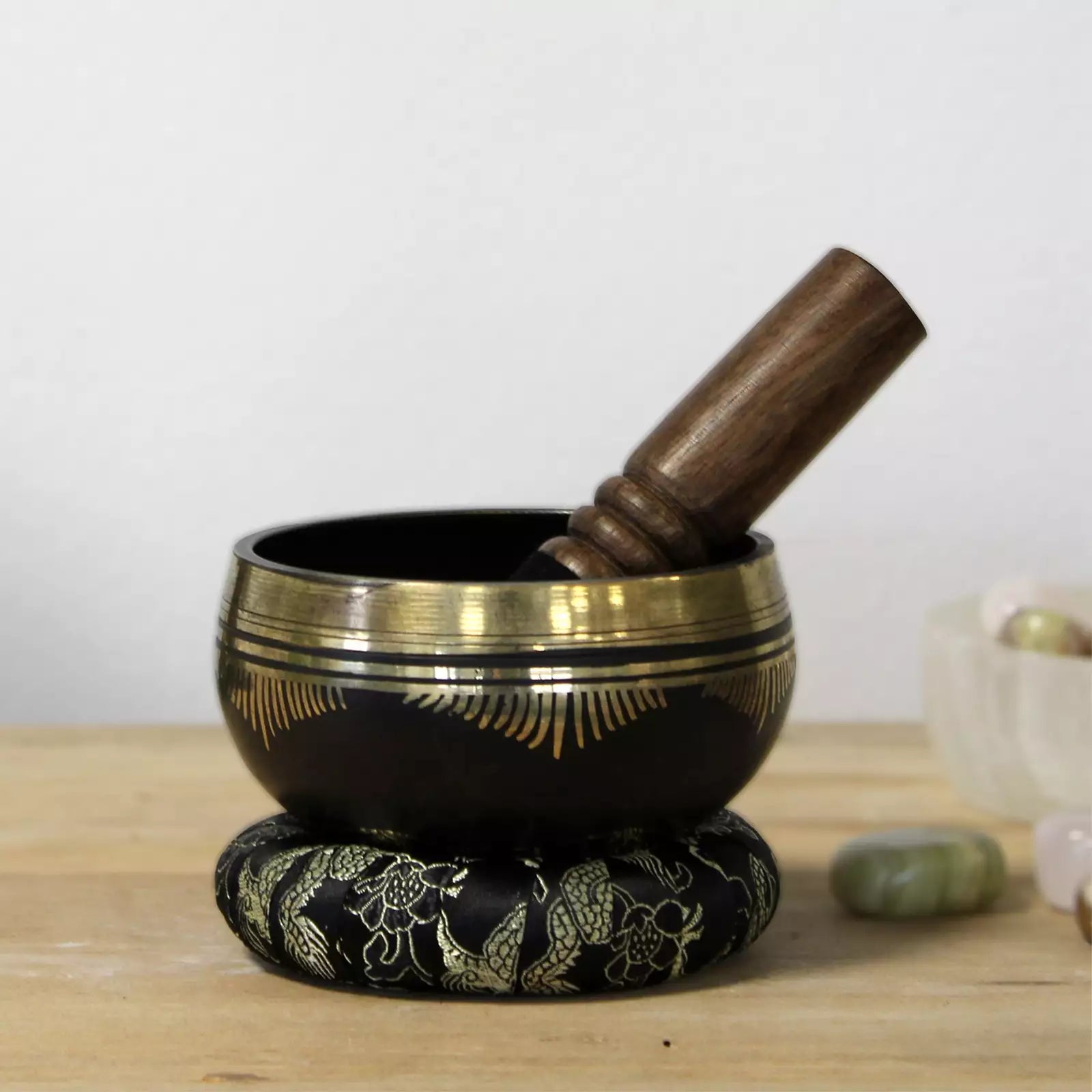 A black singing bowl with gold decorative patterns, resting on a matching black and gold decorative base, with a wooden mallet inside. The bowl is placed on a light wooden surface with a soft-focus background.