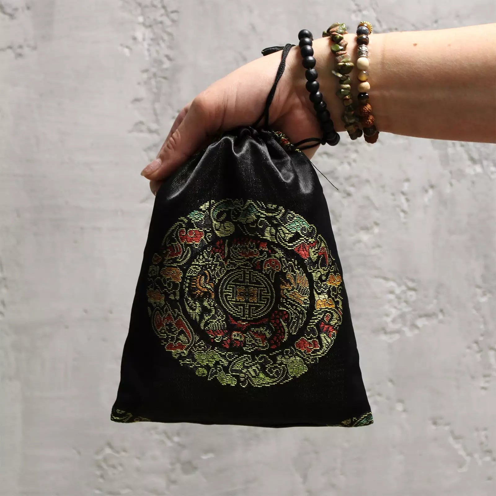 A hand holding a small black satin drawstring pouch with an ornate circular embroidered design in gold, green, red, and blue, set against a textured light gray wall.