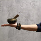 A brass singing bowl with a wooden mallet resting on an open hand, with several beaded bracelets on the wrist, set against a textured gray wall.