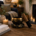 A brass singing bowl set on a wooden table in a cozy living room setting, featuring a matching wooden mallet and decorative base. The scene includes a lit candle, books, a sofa, and a fireplace in the background.