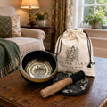 A green tara singing bowl set on a wooden table in a cozy living room. The set includes a black and gold singing bowl, a wooden mallet, a black and white patterned coaster, and a cream-colored drawstring bag with 'GREEN TARA Singing Bowl' printed on it.