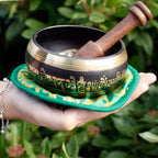A Tibetan singing bowl with gold script on a black exterior, held in a person's hand outdoors. The bowl has a wooden striker resting inside it and sits on a green fabric cushion with gold detailing, set against a background of green foliage.