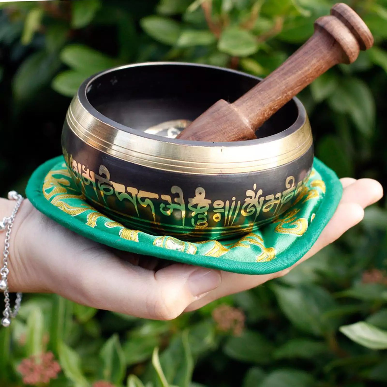 A Tibetan singing bowl with gold script on a black exterior, held in a person's hand outdoors. The bowl has a wooden striker resting inside it and sits on a green fabric cushion with gold detailing, set against a background of green foliage.