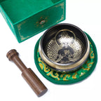 A Tibetan singing bowl set on a white background, featuring a gold-toned singing bowl with a seated deity design, a green fabric cushion with gold patterns, a wooden mallet, and a green presentation box.