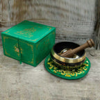A Tibetan singing bowl set with a wooden striker and a matching green decorative box, displayed on a rustic wooden surface.