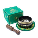 A Tibetan singing bowl set on a white background, including a black and gold singing bowl on a green decorative cushion, a wooden mallet, and a green branded box.