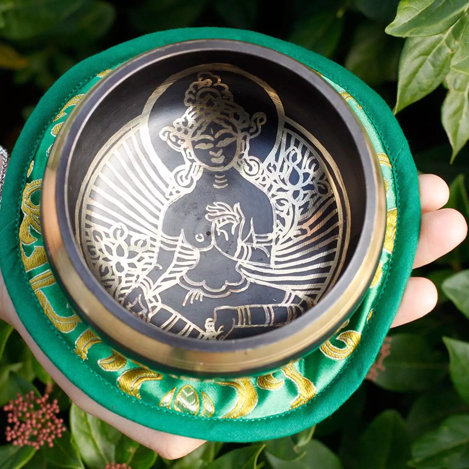 A Tibetan singing bowl with a polished bronze-toned metal interior featuring a seated Buddha figure and radiating lines, set in a round green fabric holder with gold decorative patterns. The holder is being held by a hand against a background of green leaves.