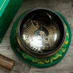 A Tibetan singing bowl with a black interior and gold-toned deity illustration, resting on a green fabric cushion with gold detailing, with a wooden mallet and part of a green box visible on a rustic wood surface.