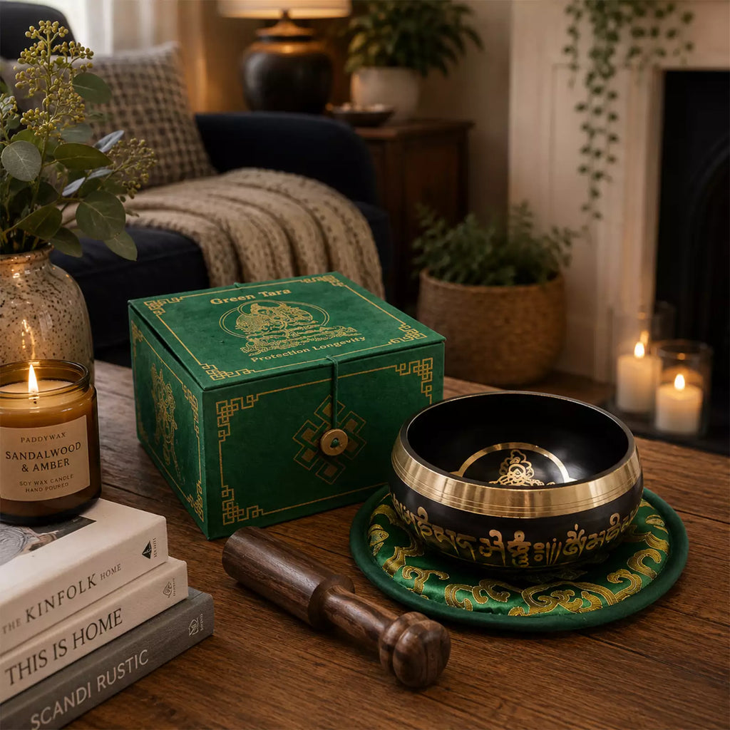 A Tibetan singing bowl set displayed on a wooden table in a cozy living room setting, next to a green decorative box, a lit candle, and stacked books. The set includes a black bowl with gold accents, a matching green and gold coaster, and a wooden mallet.