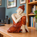 25 cm wooden thinking Buddha resting its head on its hands on a desk in a blue British study.