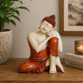 Hand Carved Wooden Buddha Statue - 25cm Thinking, featuring a serene Buddha figure with red hair and robes, seated thoughtfully on a wooden surface beside a plant and candle.
