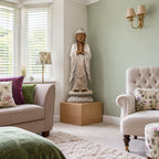 1 metre whitewashed wooden Buddha standing in prayer on an oak plinth in a British living-room corner beside a bay window.