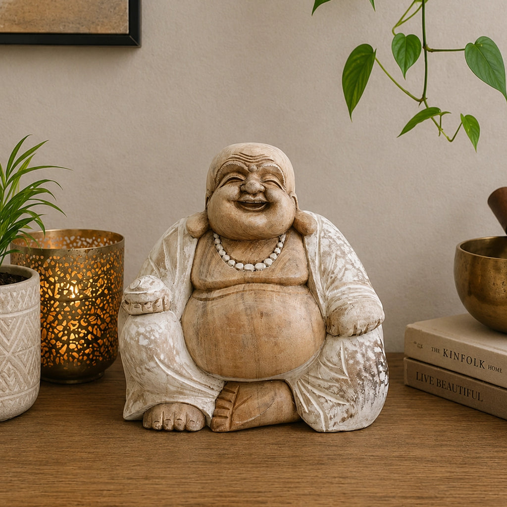 Hand Carved Wooden Happy Buddha - Whitewash 30cm sitting statue with a joyful smile, light whitewashed wood finish, displayed on a wooden surface with decorative plants and books.