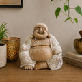 Hand Carved Wooden Happy Buddha - Whitewash 30cm sitting statue with a joyful smile, light whitewashed wood finish, displayed on a wooden surface with decorative plants and books.