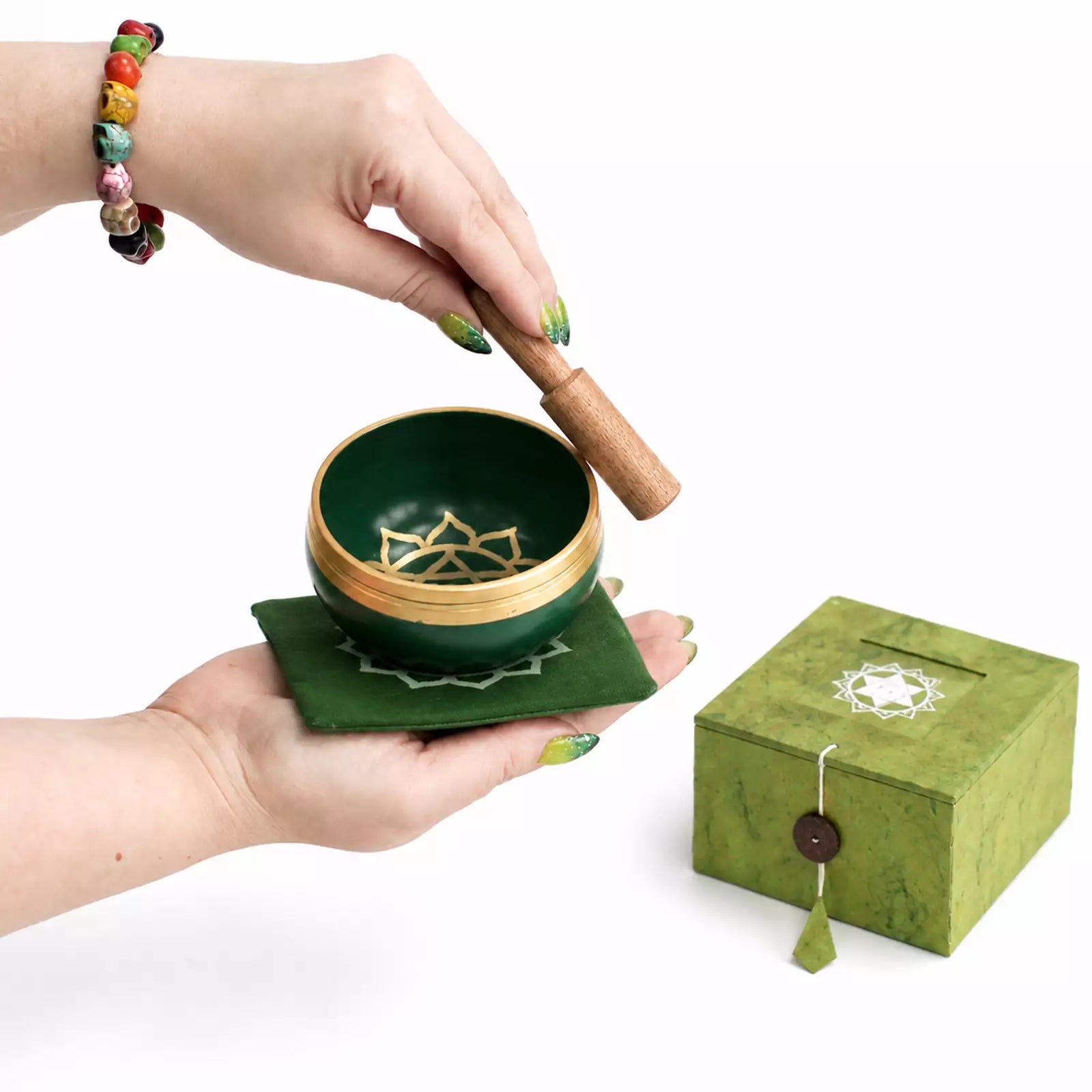 A green singing bowl with a gold-toned rim and inner star design, held by hands over a matching green cushion, with a wooden striker above it. A green box with a white star cutout and button closure sits to the right on a white background.