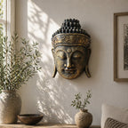 A decorative Buddha head wall sculpture in an antique bronze finish with gold accents, mounted on a warm beige wall in a sunlit room with potted plants and a wooden table below.