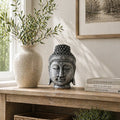 A decorative Buddha head statue in a weathered silver finish sits on a wooden console table next to a speckled ceramic vase with greenery and a stack of books.