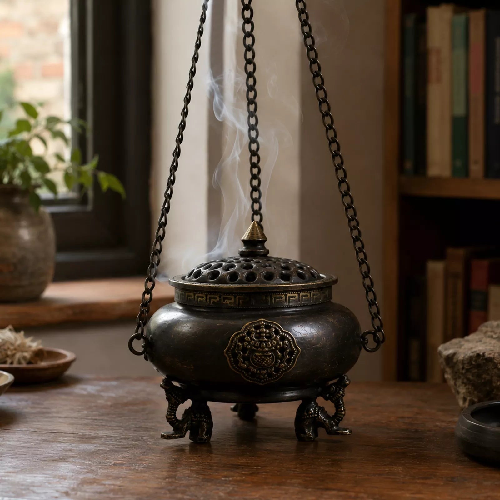 Large Brass Tibetan Burner - Four Symbol Hanging Pot with intricate designs and ornate legs, gently releasing smoke, displayed on a wooden surface in a cozy room setting.