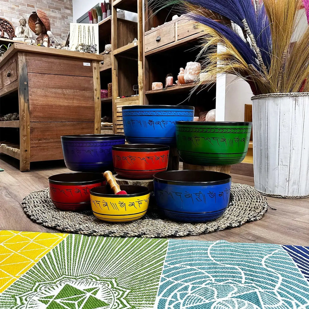 Large Chakra Set of Seven Singing Bowls (19.5-35cm) arranged on woven mat in vibrant colors with wooden shelves and decorative items in the background.