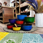 Large Chakra Set of Seven Singing Bowls (19.5-35cm) arranged on woven mat in vibrant colors with wooden shelves and decorative items in the background.