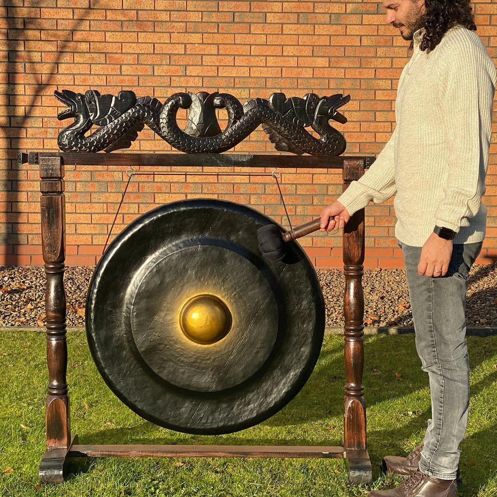 Person striking a large gong on a wooden stand outdoors against a brick wall.