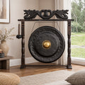 A large round black gong with a polished gold center, mounted on a dark wooden stand with dragon carvings at the top, shown in a styled room setting.