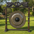 A large decorative outdoor gong with an ornate wooden stand, featuring a dragon motif at the top and a hanging mallet, set on a green lawn with trees in the background.