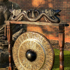 A large round bronze gong with a carved wooden frame and dragon headpiece, photographed outdoors in front of a brick wall and stone statue.